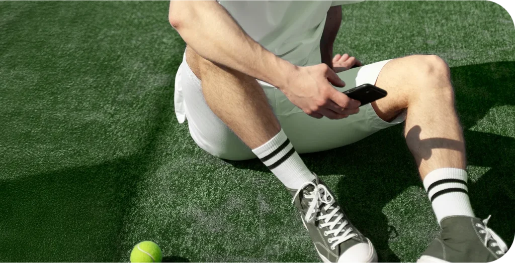 Egy padel játékos ül a padel pályán telefonnal a kezében egy labda mellett